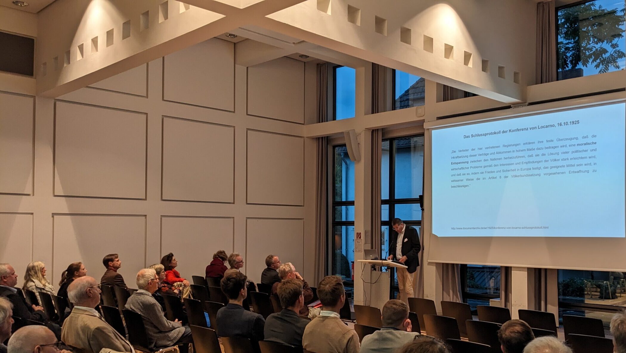 Zuhörer und Vortragender vor einer Leinwand mit Vortragstext.